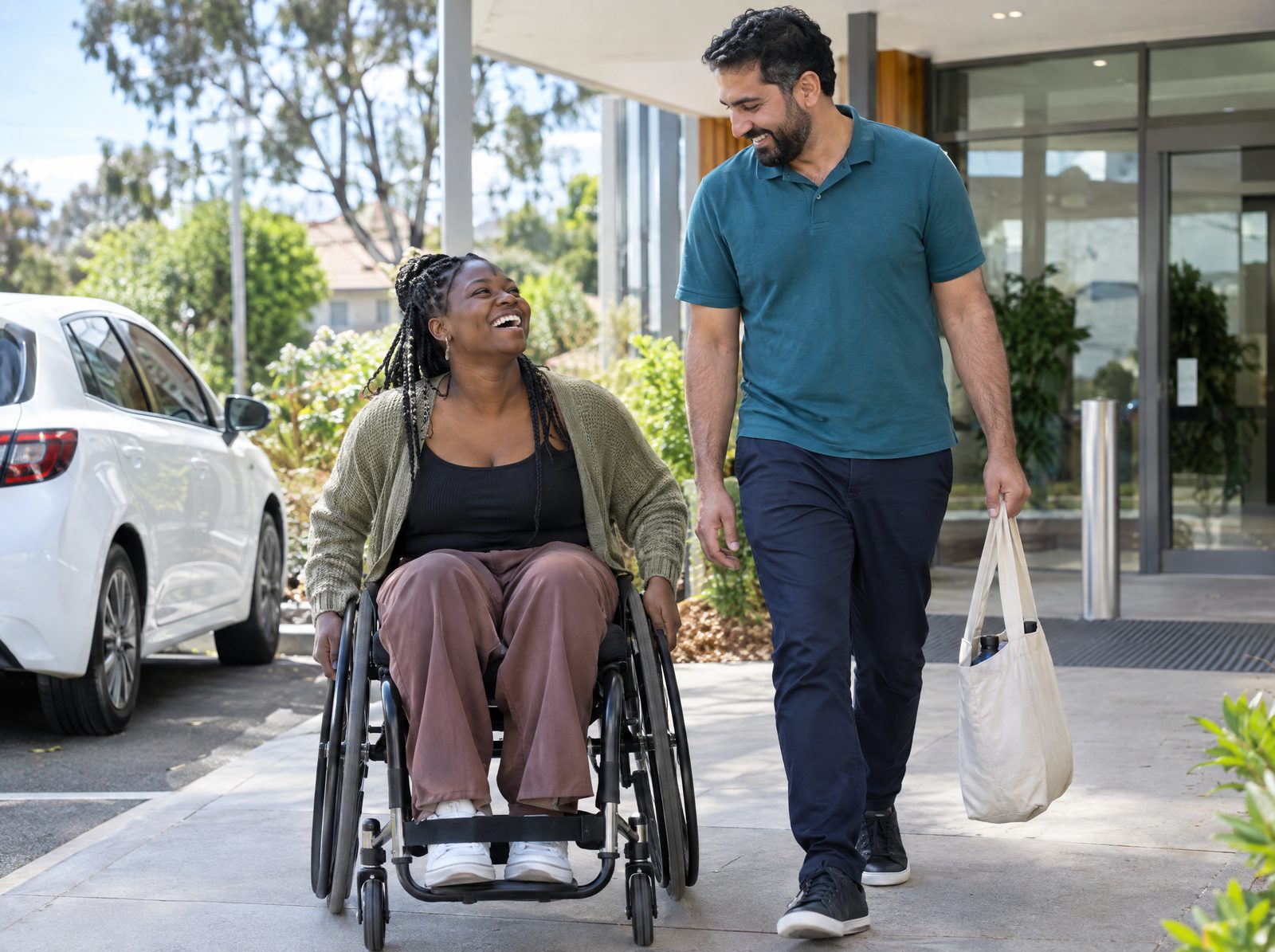 A young woman in a wheelchair laughs with her support worker as they arrive at a health clinic